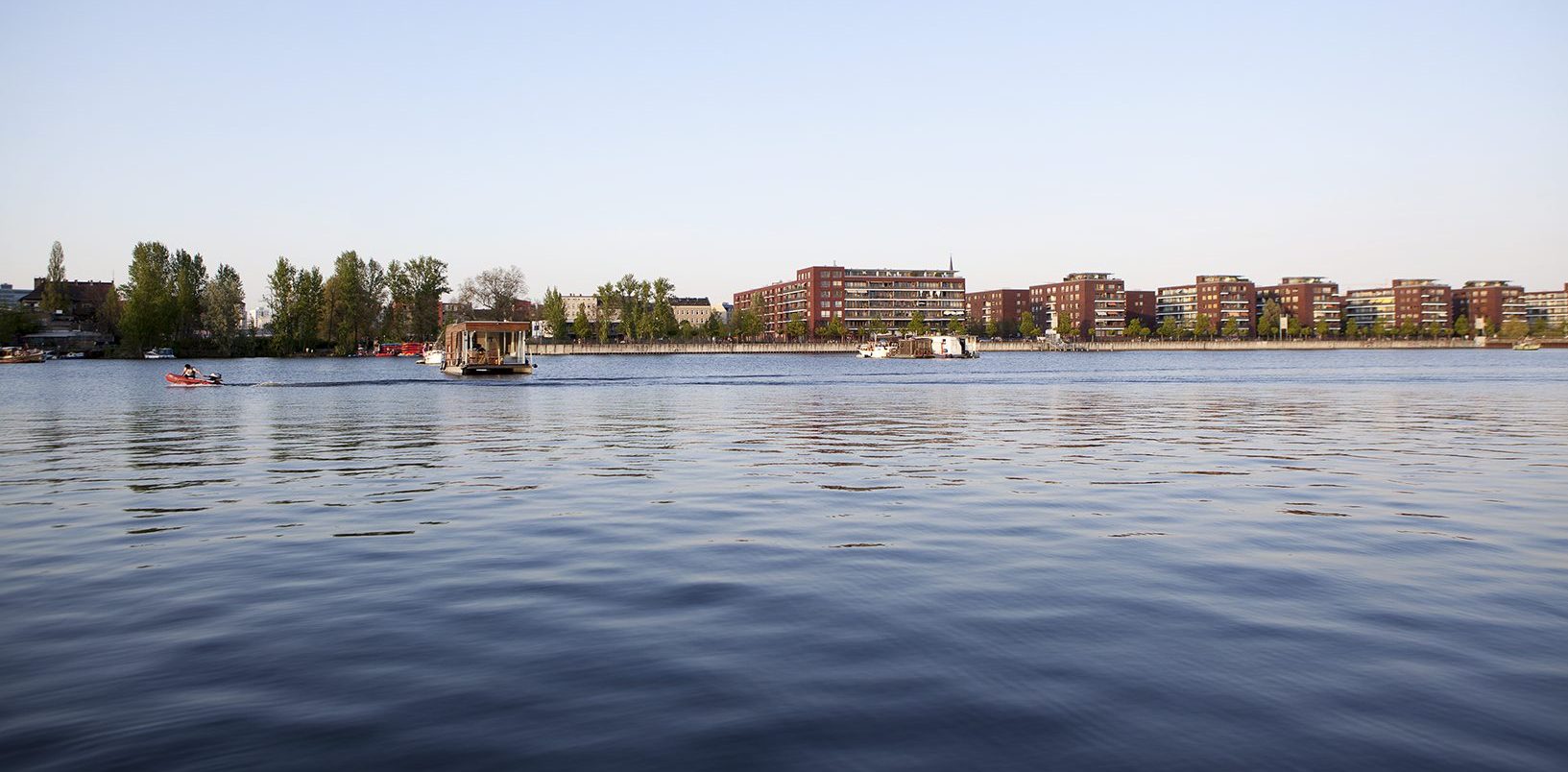 Man sieht einen großen See oder Fluss. Auf dem Wasser fahren Boote. Hinten am Ufer stehen Häuser und viele Bäume.
