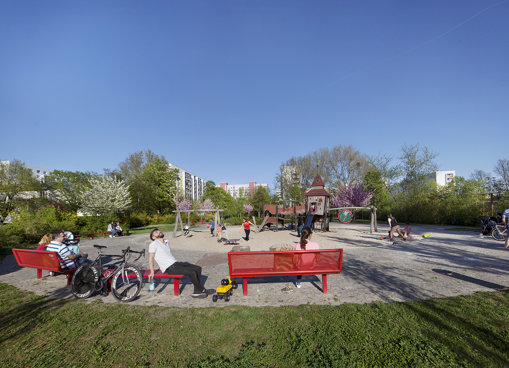 Man erkennt einen Spielplatz inmitten einer Wohnsiedlung. Der Spielplatz ist umrahmt von Bäumen. Dahinter liegen Hochhäuser. Auf dem Spielplatz spielen fröhliche Kinder. Auf den roten Bänken im Vordergrund entspannen Erwachsene. Der Himmel ist blau und die Sonne scheint.