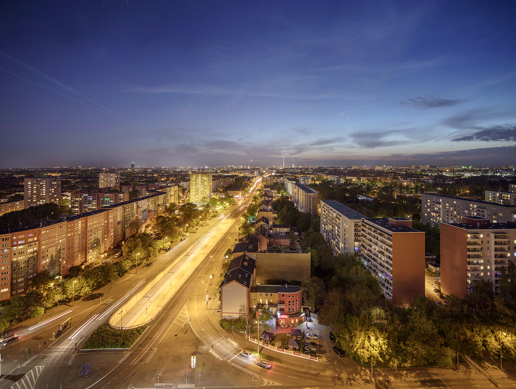 Das Bild zeigt eine große Straße mit vielen Häusern. Es ist abends, die Straße ist hell beleuchtet. Am Himmel sieht man ein paar Wolken.