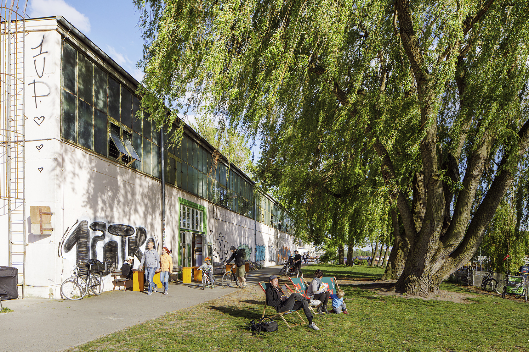 Mehrere Menschen sitzen auf Stühlen und Bänken vor einem Gebäude mit Graffiti. Rechts neben dem Gebäude steht ein großer Baum mit vielen grünen Blättern. Die Sonne scheint.