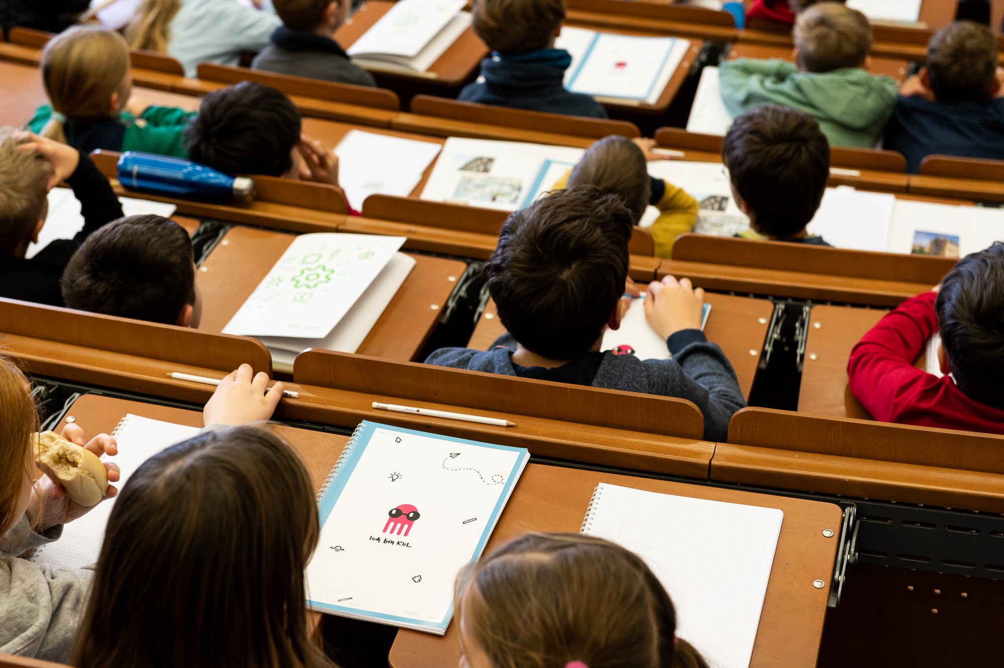 Viele Kinder sitzen in einem Hörsaal. Sie schauen nach vorn und haben Hefte und Stifte vor sich auf dem Tisch.