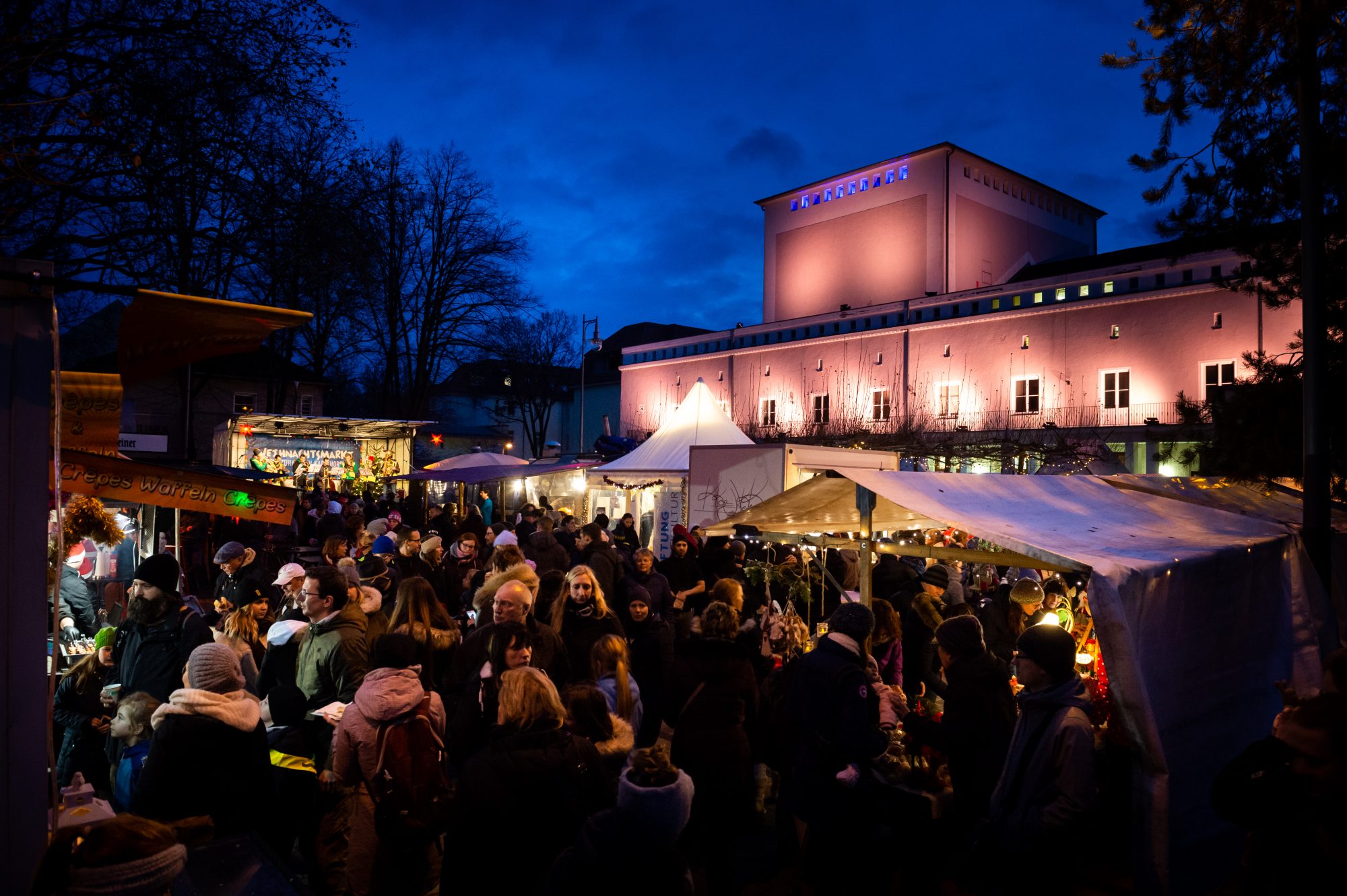 Abgebildet sind viele Menschen, die einen Markt am Abend besuchen. Es gibt viele bunte Lichter und Verkaufsstände. Im Hintergrund ist ein großes, pink beleuchtetes Gebäude.