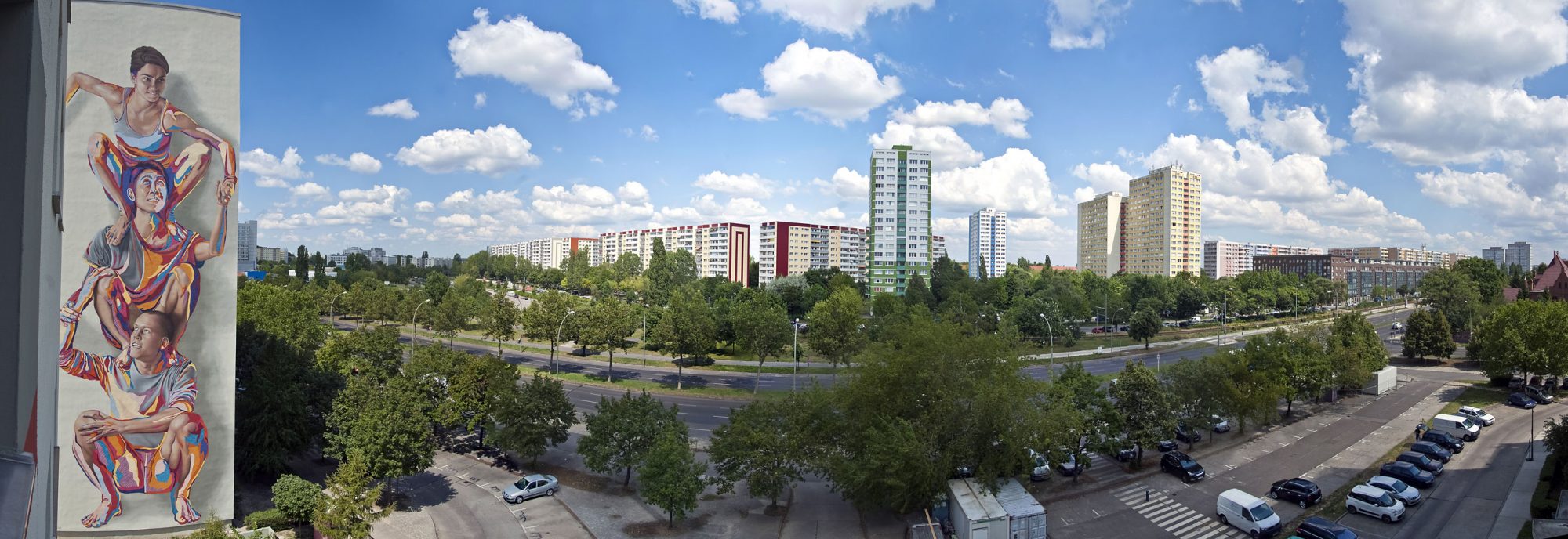 Man sieht eine Panorama Aufnahme der Stadt mit vielen Bäumen, Straßen und hohen Häusern. Links ist ein buntes Graffiti Wandbild mit drei Menschen aufeinander hockend. Der Himmel ist blau mit vielen vielen Wolken.