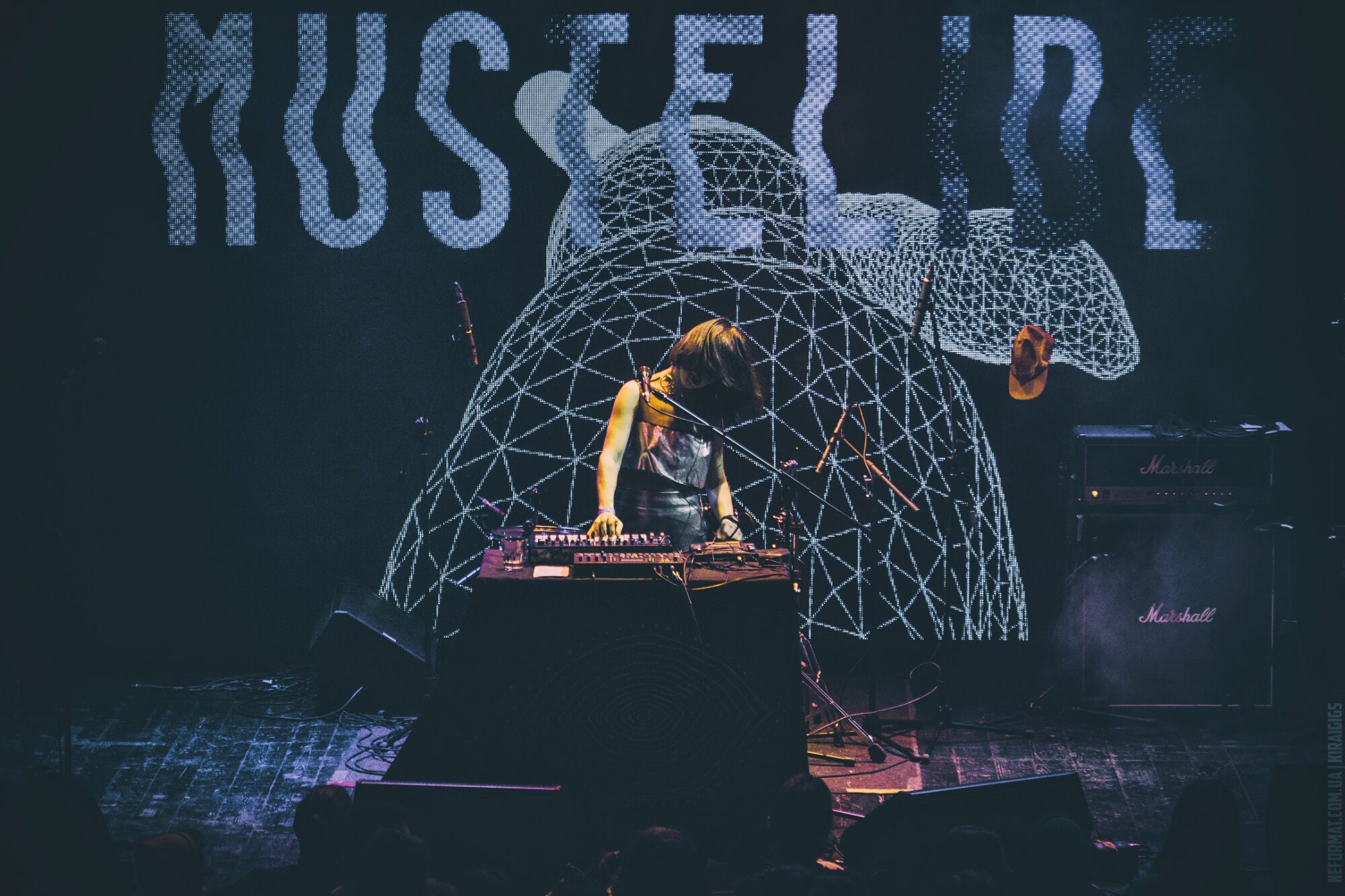 Eine Person steht hinter einem DJ- Pult auf der Bühne. Hinter ihr leuchten bunte Lichter und ein großes Bild mit Schrift.
