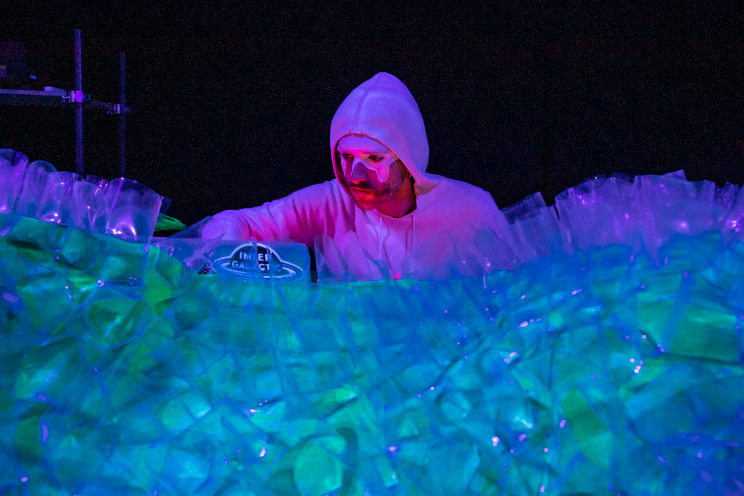Eine Person in einem weißen Hoodie steht hinter vielen leuchtenden Plastiktüten. Die Farben sind blau, grün und lila. Es handelt sich um eine Bühnen-Show.