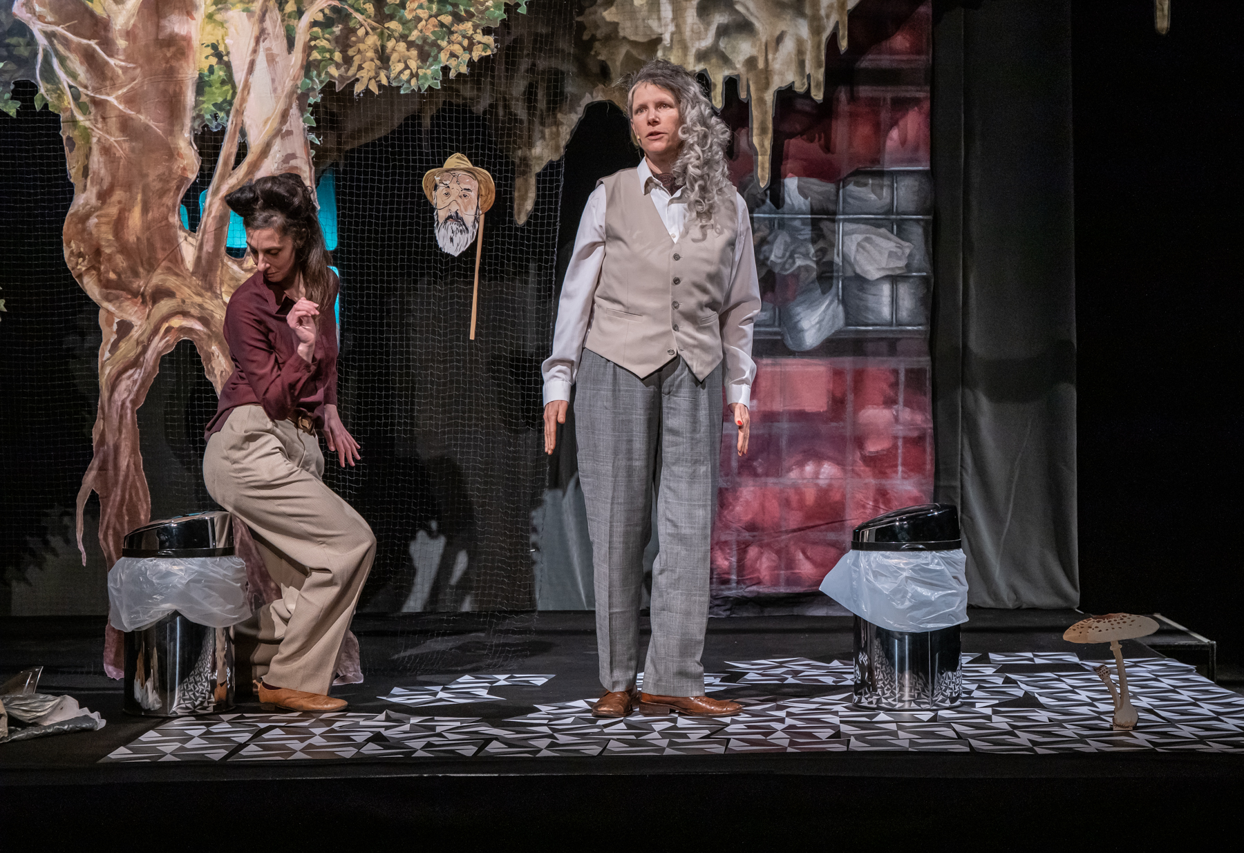 Zwei Menschen stehen auf einer Theaterbühne. Hinter ihnen ist ein gemalter Baum und eine bunte Kulisse. Beide tragen Kostüme und schauen ernst.