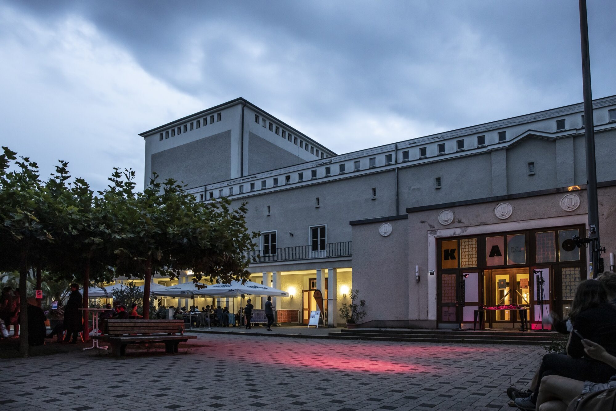 Abgebildet ist das KAHO Theater am Abend mit beleuchteten Fenstern. Vor dem Haus ist ein Platz mit Bäumen und Sitzbänken. Der Himmel ist bewölkt.