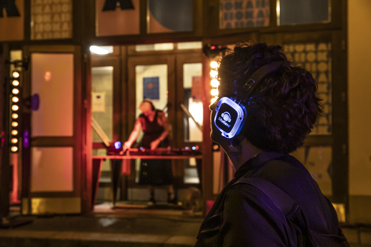 Eine Person trägt leuchtende Kopfhörer und schaut auf einen DJ, der vor einem Gebäude Musik macht. Es ist Abend, und bunte Lichter leuchten.