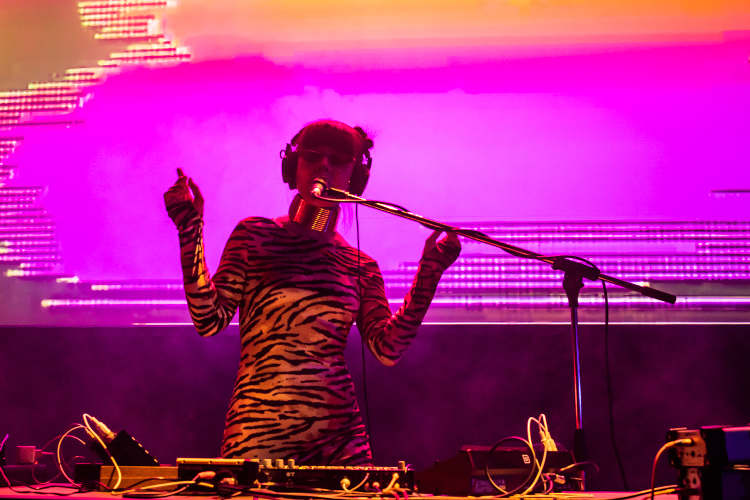 Eine Frau spielt Musik auf einer Bühne. Sie trägt ein Zebra-Kleid und hat Kopfhörer auf. Hinter ihr leuchten bunte Lichter in Pink und Orange.