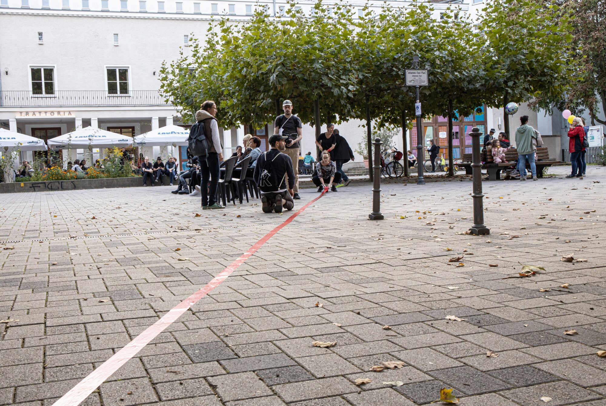 Mehrere Menschen sitzen auf dem Platz vor dem KAHO Theater. Sie schauen nach vorn. Eine rote Linie verläuft über den Boden. Im Hintergrund sind Bäume, Cafétische und ein weißes Gebäude.