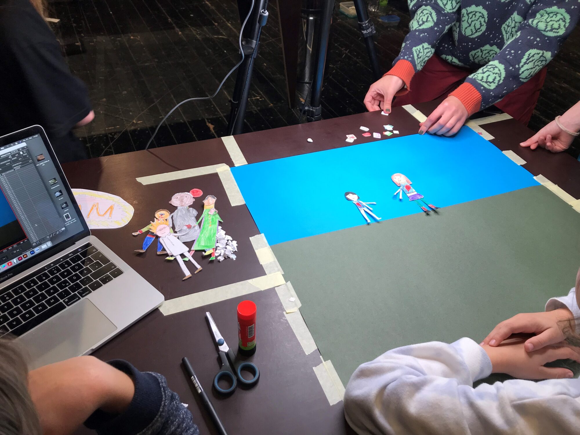 Mehrere Kinder basteln an einem Tisch. Auf buntem Papier liegen ausgeschnittene Figuren. Ein Laptop steht daneben.