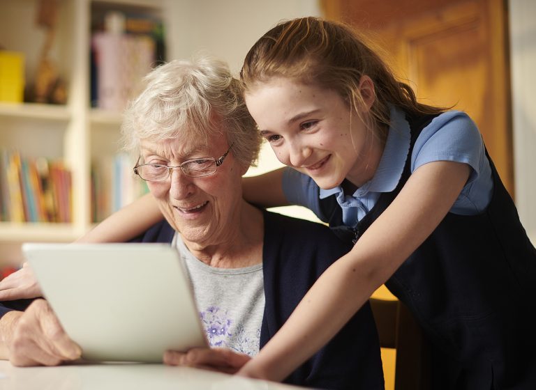 Eine ältere Dame und ein jüngeres Mädchen schauen gemeinsam auf ein Tablet. Beide lächeln. Sie sitzen an einem Tisch.