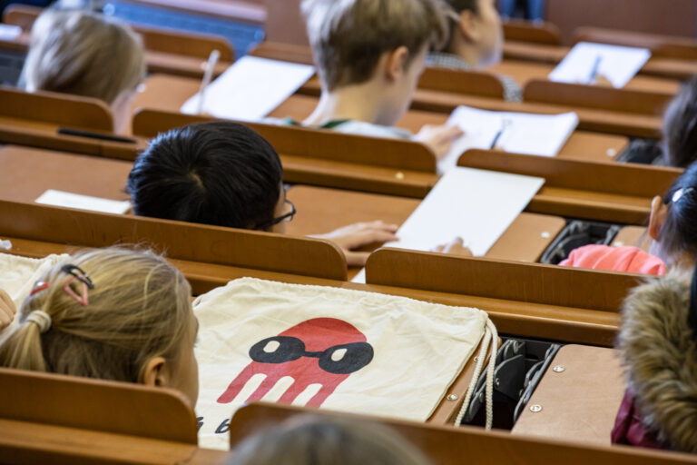 Mehrere Kinder sitzen in einem Hörsaal und schreiben auf Papier. Im Vordergrund liegt ein Stoffbeutel mit dem Motiv eines roten Tintenfischs mit schwarzen Augen und Maske auf einem Klapptisch.