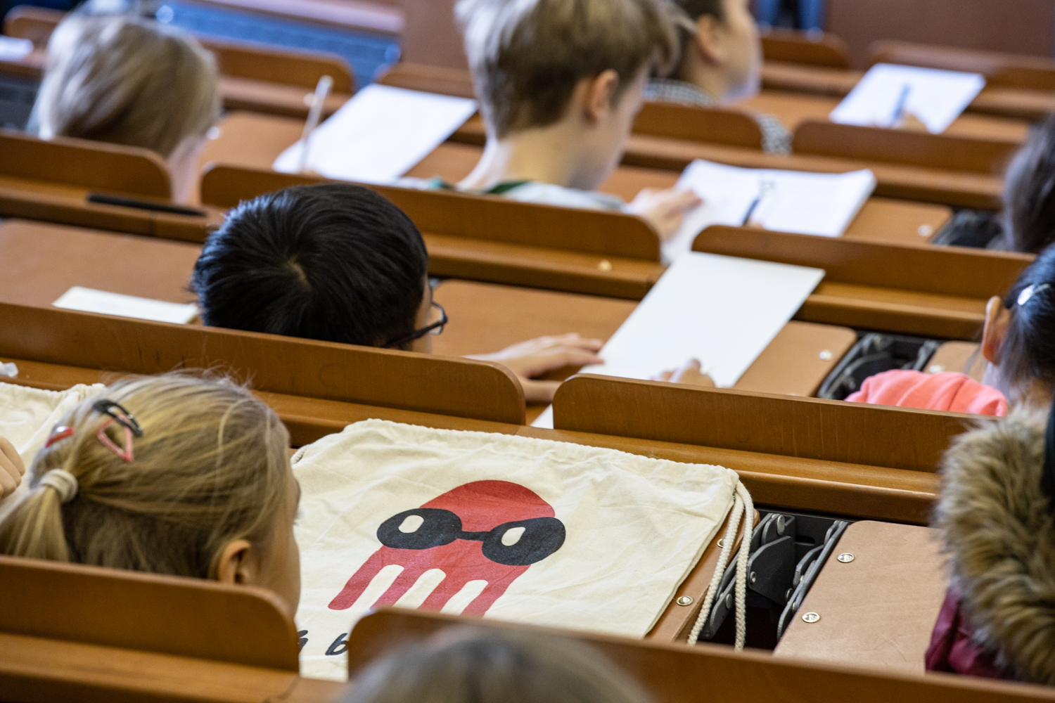 Mehrere Kinder sitzen in einem Hörsaal und schreiben auf Papier. Im Vordergrund liegt ein Stoffbeutel mit dem Motiv eines roten Tintenfischs mit schwarzen Augen und Maske auf einem Klapptisch.
