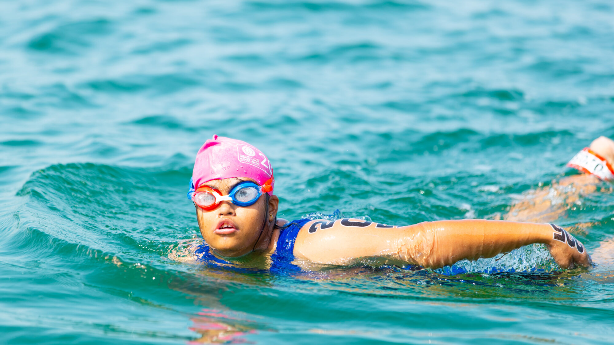 Ein junges Mädchen mit geistiger Behinderung schwimmt im offenen Wasser. Sie trägt eine pinke Badekappe, Schwimmbrille und einen blauen Badeanzug. Eine Startnummer ist auf ihrem Oberarm sichtbar. Sie nimmt konzentriert an einem Wettkampf teil.