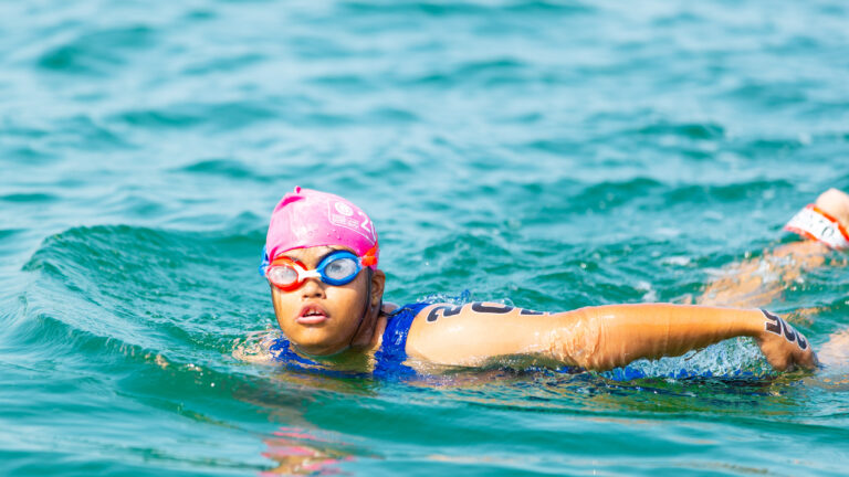 Ein junges Mädchen mit geistiger Behinderung schwimmt im offenen Wasser. Sie trägt eine pinke Badekappe, Schwimmbrille und einen blauen Badeanzug. Eine Startnummer ist auf ihrem Oberarm sichtbar. Sie nimmt konzentriert an einem Wettkampf teil.
