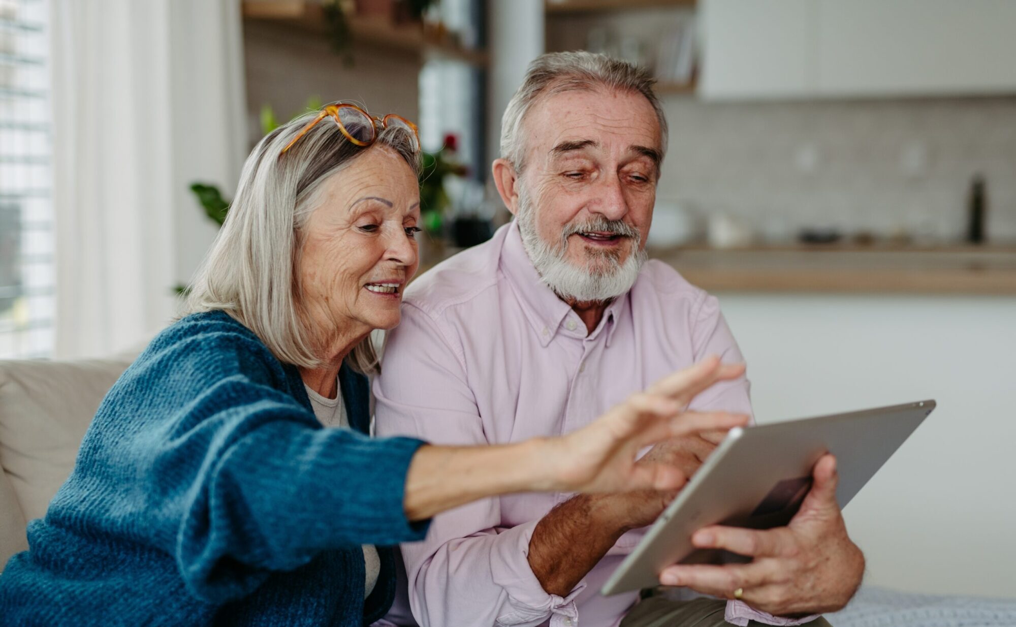 Eine Seniorin und ein Senior bedienen gemeinsam ein digitales Tablet. Sie sitzen in einer Wohnung.