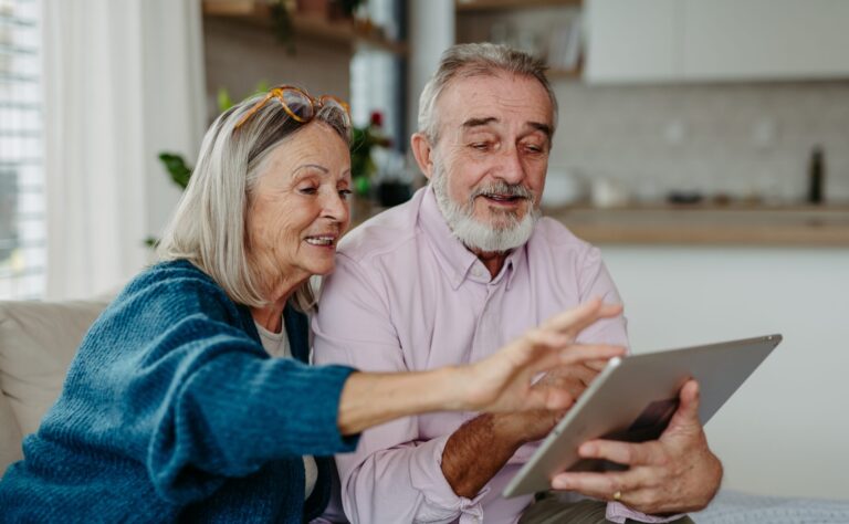Eine Seniorin und ein Senior bedienen gemeinsam ein digitales Tablet. Sie sitzen in einer Wohnung.