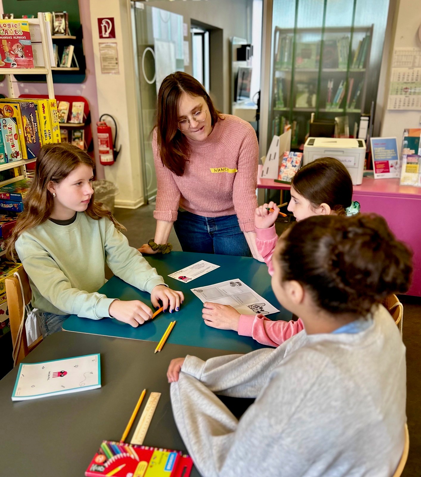 Drei Kinder sitzen um einen Tisch und unterhalten sich. Vor ihnen liegen Stifte und Arbeitsblätter. Eine Erwachsene steht am Tisch und hört den Kindern zu. Im Hintergrund ist eine Bibliothek zu sehen.
